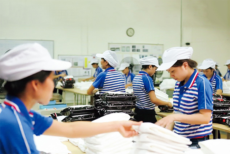 白いキャップと横縞ポロシャツを着た複数人が、作業台で白い布製品をたたんだり、包装された製品を扱ったりしている。