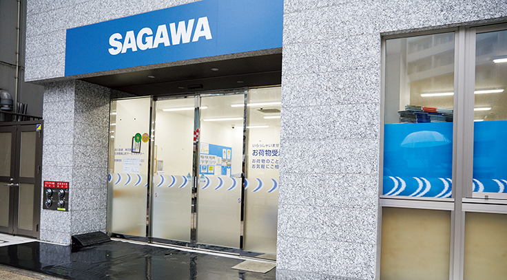 SAGAWAの青い看板がある建物の入り口。ガラスの自動ドアに案内文字があり、左側の壁面に消火栓がある。右側の窓には青いシートが貼られている。