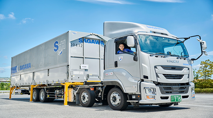 停車している大型セミトレーラー。運転席では人物が窓から顔を覗かせている。シルバーの車体にはSAGAWAやSGHのロゴが記されている。