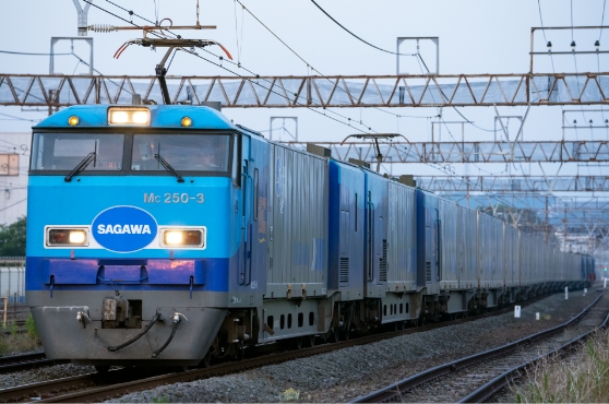 SAGAWAのロゴとMc 250-3が記された青い電気機関車が、貨物コンテナ車両を牽引し線路上を走る。