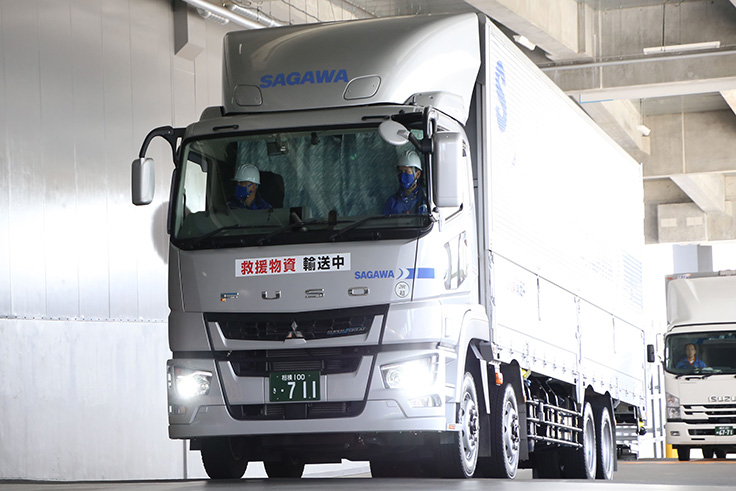 SAGAWAの大型トラック。救援物資輸送中の表示があり、2人の人物が乗車している。