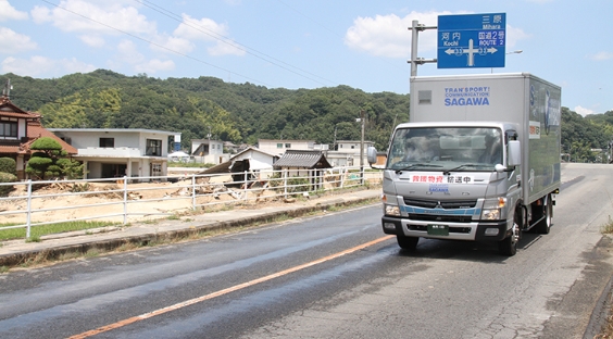 救援物資輸送中と書かれたSAGAWAのトラックが、倒壊した建物横の道路を走る様子。