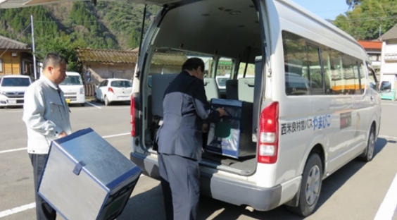 西米良村バスやまびこの白いバン後部に、二人の人物が荷物を積み込む。一人が箱を持ち、もう一人が荷台に載せている。
