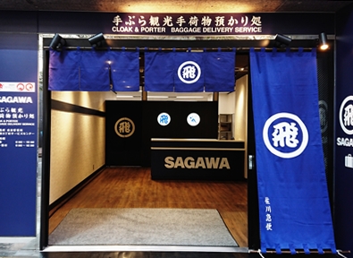 手ぶら観光手荷物預かり処の店舗入口。上部の看板には CLOAK and PORTER BAGGAGE DELIVERY SERVICE と英語で書かれている。入口には飛のマークが入った青い暖簾が下がり、右側の長い暖簾には佐川急便の文字がある。