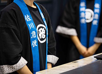 佐川急便の文字が入った青い襟の黒い法被を着た二人の人物。胸元には丸の中に飛の文字が入った白いロゴがある。