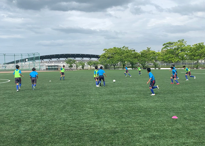 人工芝のサッカーフィールドで練習をする複数の人物。青いTシャツと紺色のハーフパンツを着用し、一部の選手は蛍光イエローのビブスを着ている。緑の木々とスタジアムを背景に、白いボールとピンクのマーカーコーンが置かれている。