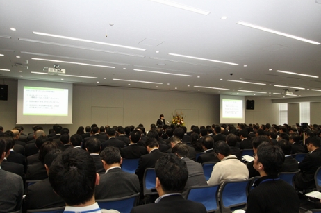 多数の参加者が会議室で講演を聞く。壇上の講演者が資料をスクリーンに投影。