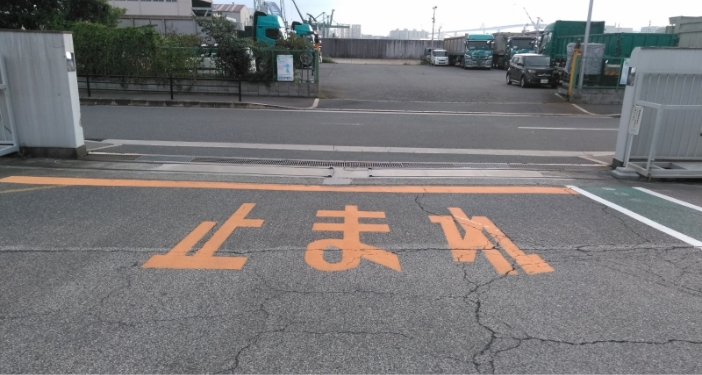 オレンジ色の停止線と 止まれ の文字がある路面。その先の道路を挟んで、奥にトラックが並ぶ駐車場がある。