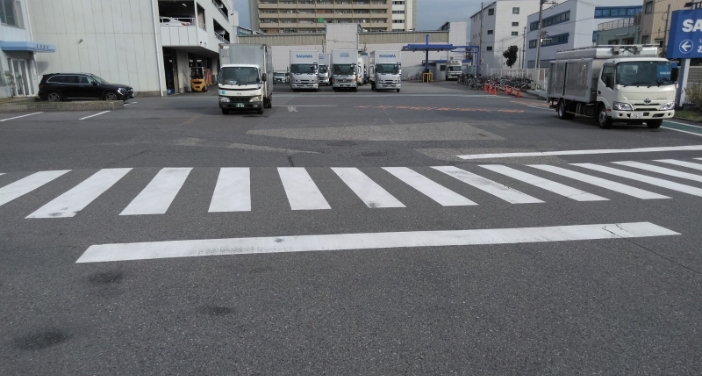 手前に横断歩道と停止線が描かれたアスファルトの路面。奥には複数の運送トラックが並ぶ建物と駐車場がある。