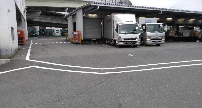 二本の白い線が引かれた路面と、その奥の屋根付き荷捌き場に停車している二台の大型トラック。