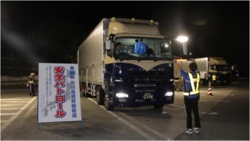 大型トラックと 安全パトロール の看板があり、反射ベストの人物が誘導する。