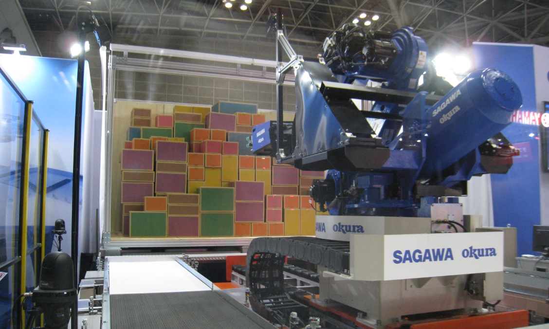 屋内施設に設置された、SAGAWA okuraのロゴが入った青い大型ロボット。手前にはベルトコンベアがあり、奥には色やサイズが異なる様々な箱が壁状に高く積み上げられている。