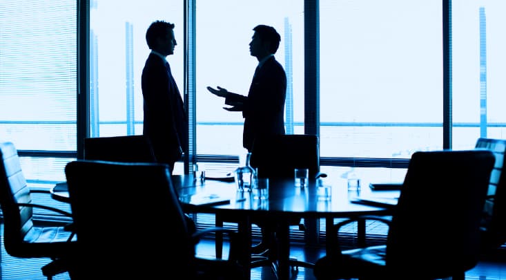 窓際でスーツ姿の2人が円形テーブルを囲む。テーブルにはノートPCとグラスがある。