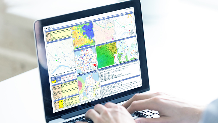 ラップトップPCの画面に、地図やグラフなどの複数のデータパネルが表示されている様子。手前でにはキーボードを操作する両手が写っている。