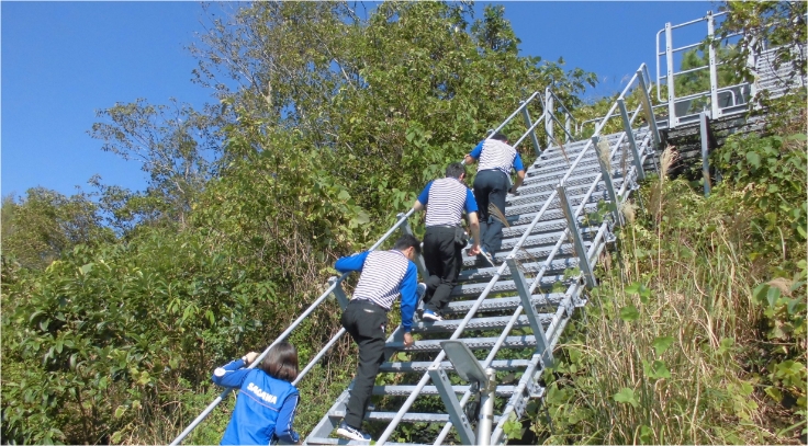 背中に「SAGAWA」の文字がある人物を含む数人が金属製の階段を上る。