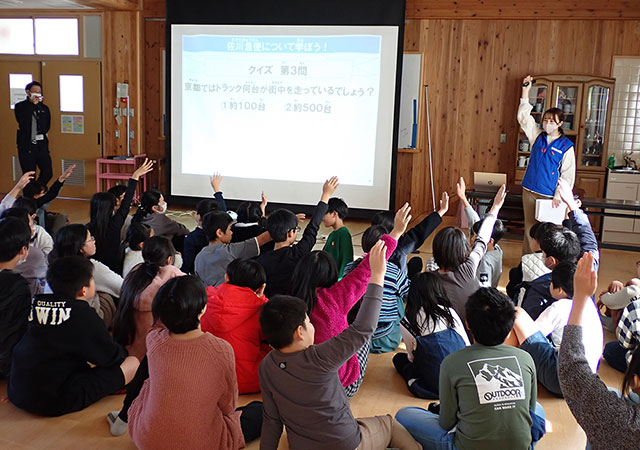 佐川急便について学ぼう！のスライドが投影された教室で、多くの子供たちがクイズに挙手。スライドには クイズ 第3問 京都ではトラック何台が街中を走っているでしょう？ 1 約100台 2 約500台 とある。