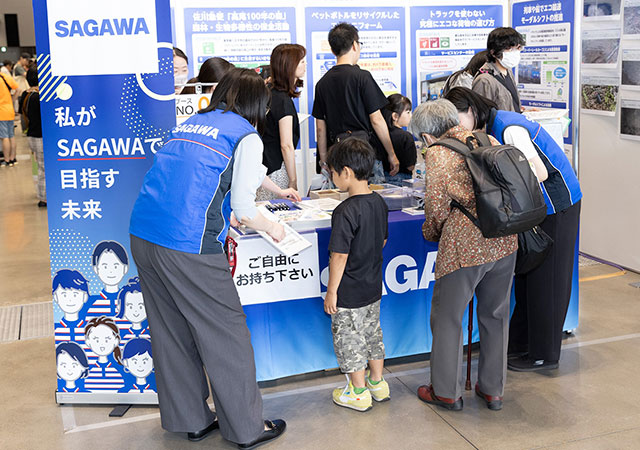 SAGAWAのブースで人々が 私がSAGAWAで目指す未来 の展示を見ている。森林・生物多様性の保全活動や、ペットボトルリサイクル、エコ輸送について説明されている。