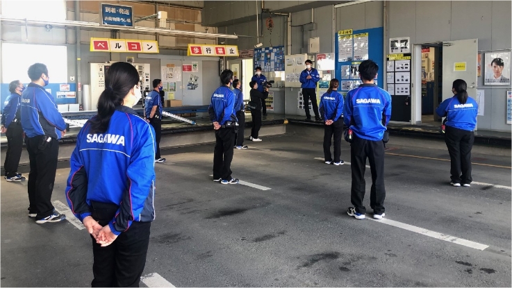 SAGAWAの制服を着た複数の人物が、屋内の作業場で床の印に沿って並んでいる様子。
