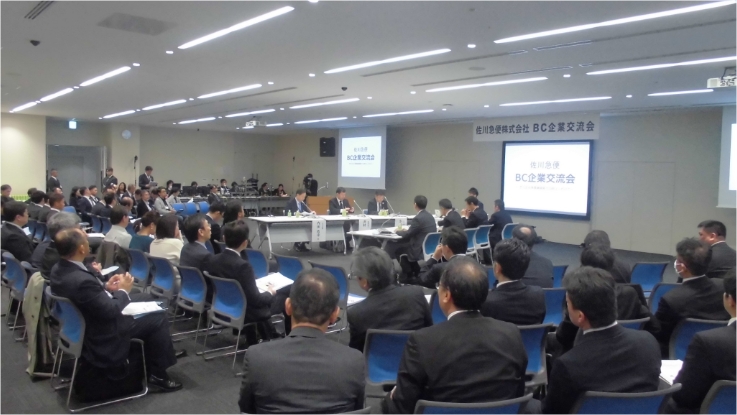 佐川急便のBC企業交流会の会場風景。壇上に数名の登壇者が座り、前方のスクリーンには会合の名称が表示されている。客席には多数の参加者が着席している。