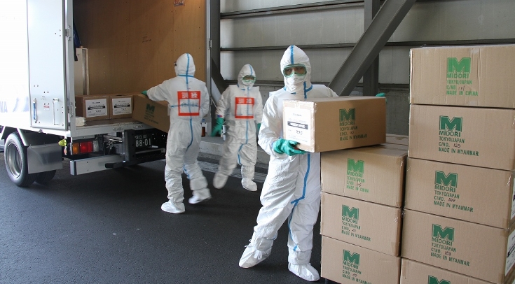 防護服姿の3人の作業員がトラックから段ボール箱を運び出す様子。作業員の背中には文字の入ったオレンジ色のゼッケンがあり、箱にはMIDORI TOKYO, JAPANのロゴやMADE IN CHINAといった文字が記されている。