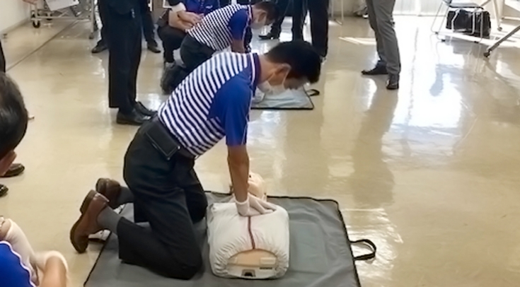 青と白のボーダー柄のシャツを着て、白いマスクを着用した人物たちが、一列に並んでダミー人形に胸骨圧迫を行う様子。心肺蘇生の訓練を受けている。