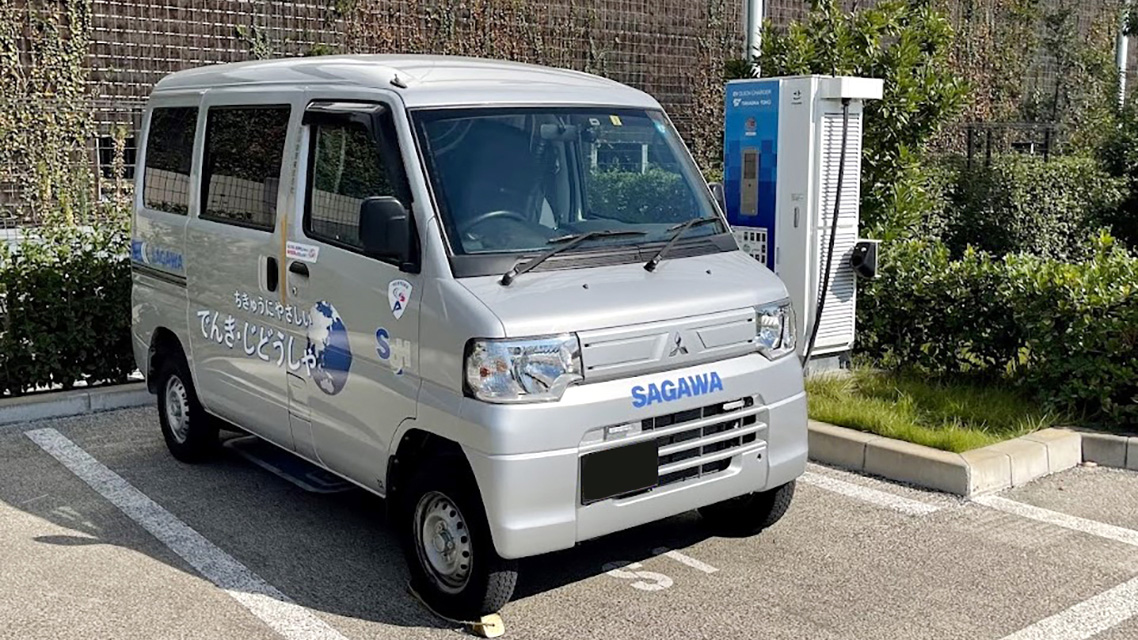 駐車場で充電中のシルバーの小型電気自動車。車両の側面には地球のイラストとちきゅうにやさしいでんきじどうしゃという文字があり、隣には充電器が設置されている。