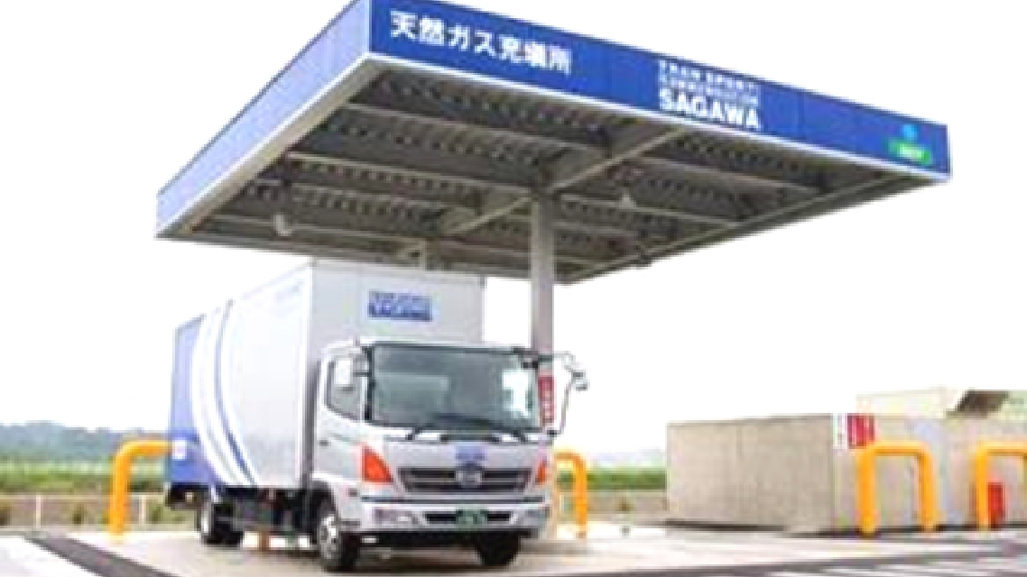 天然ガス充填所の青い屋根の下に停車している大型トラック。周囲には充填設備や黄色のガードパイプが設置されている。