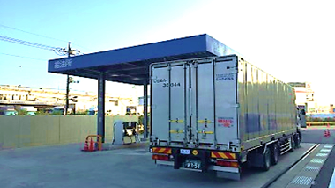 青い屋根の給油所に停車している大型トラックの背面。荷台の扉には識別番号やロゴが描かれており、周囲には給油設備がある。