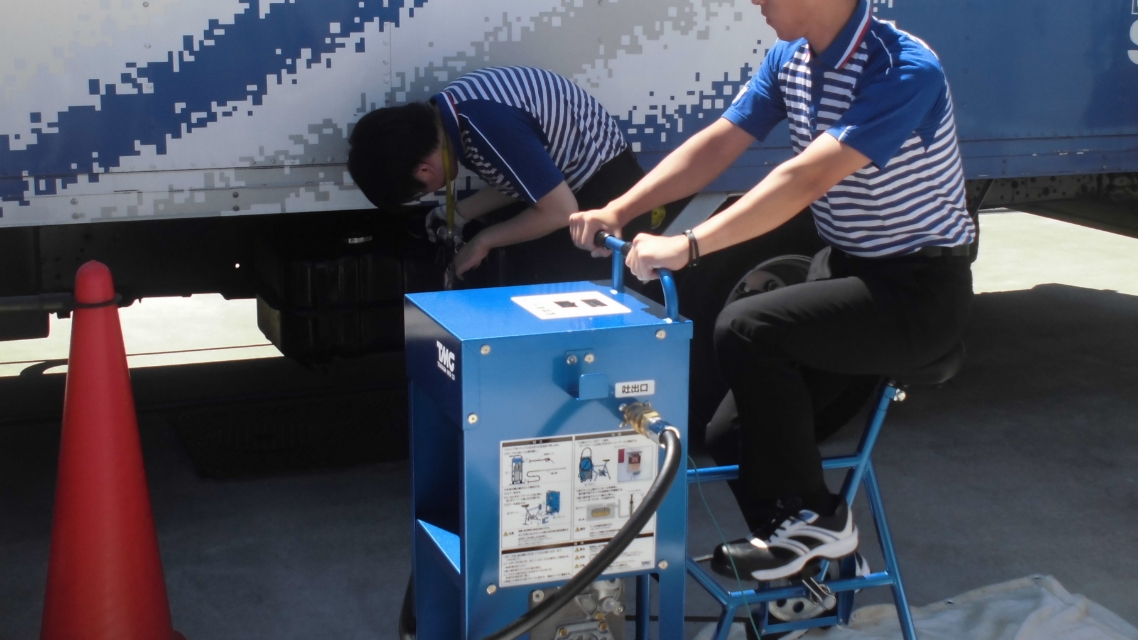 トラックのそばで青い装置を操作している二人の作業員。一人は手前の装置にまたがってハンドルを握り、もう一人は奥でトラックの車体下を点検している。