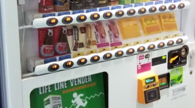 飲料や食品が並ぶ災害対応型の自動販売機。下部にLIFE LINE VENDERと書かれており、価格表示のランプが点灯している。